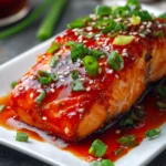 Close-up of tender baked salmon fillet centered on a white rectangular plate, topped with sticky bright red-orange sweet chili garlic sauce, garnished with chopped green onions and white sesame seeds on a rustic surface.