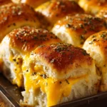 Close-up of golden baked turkey and cheese sliders in a rectangular baking dish, showing melted cheddar cheese between soft slider buns and sprinkled with herbs.