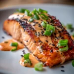 Close-up of pan-seared salmon fillet with crispy exterior and tender flaky interior on a white plate, drizzled with creamy spicy bang bang sauce, garnished with sliced green onions and black sesame seeds