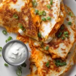 Close-up overhead photo of crispy buffalo chicken quesadillas cut into wedges with melted cheddar and mozzarella cheese, vibrant buffalo sauce, green onion garnish, and ranch dressing on a white plate with warm natural lighting.