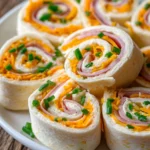Close-up of cream cheese roll-ups sliced into pinwheels arranged on a white plate, highlighting layers of cream cheese with herbs, sliced deli ham, shredded cheddar cheese, and chopped fresh chives wrapped in soft flour tortillas.