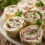 Close-up of sliced cream cheese roll-ups with herbs and deli ham arranged in a circular pattern on a white plate, showing soft tortillas and visible green herb specks.