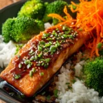 Close-up of a crispy pan-seared salmon fillet with golden skin on white jasmine rice, steamed broccoli florets, shredded carrots, drizzled with soy-ginger glaze and garnished with sesame seeds and chopped scallions on a rustic wooden surface