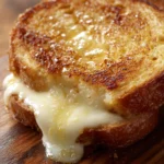 Close-up of a grilled mozzarella sandwich cut diagonally, showing golden crispy bread and melted mozzarella cheese with a buttery sheen on a white plate