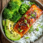 Close-up of a honey soy glazed pan-seared salmon fillet served on white jasmine rice with steamed broccoli florets and sliced avocado, garnished with sesame seeds and chopped green onions on a rustic wooden surface in warm natural light.