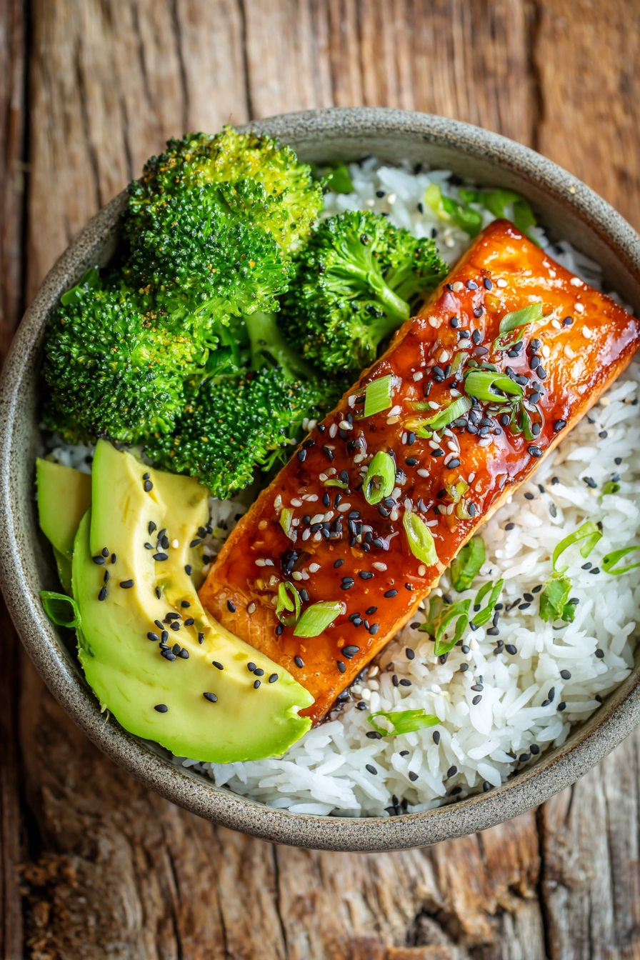 Close-up of a honey soy glazed pan-seared salmon fillet served on white jasmine rice with steamed broccoli florets and sliced avocado, garnished with sesame seeds and chopped green onions on a rustic wooden surface in warm natural light.