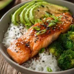Close-up of a honey glazed salmon fillet with crispy edges on a bed of white jasmine rice, steamed broccoli florets and sliced avocado arranged neatly, garnished with black sesame seeds and chopped green onions on rustic wooden surface under warm natural light.