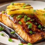 Close-up of crispy pan-seared salmon fillet with glossy pineapple glaze, grilled pineapple slice, and chopped green onions on a white plate with warm natural light