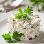Close-up of classic tuna salad neatly molded on a white plate, creamy tuna mixed with mayonnaise, lemon juice, Dijon mustard, finely chopped celery and red onion, garnished with a small parsley sprig, presented in a clean and minimalistic style.