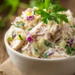 Close-up of classic tuna salad in a shallow white bowl showing creamy texture with chunks of tuna, finely chopped celery and red onion, topped with a sprig of bright green parsley on a rustic wooden surface