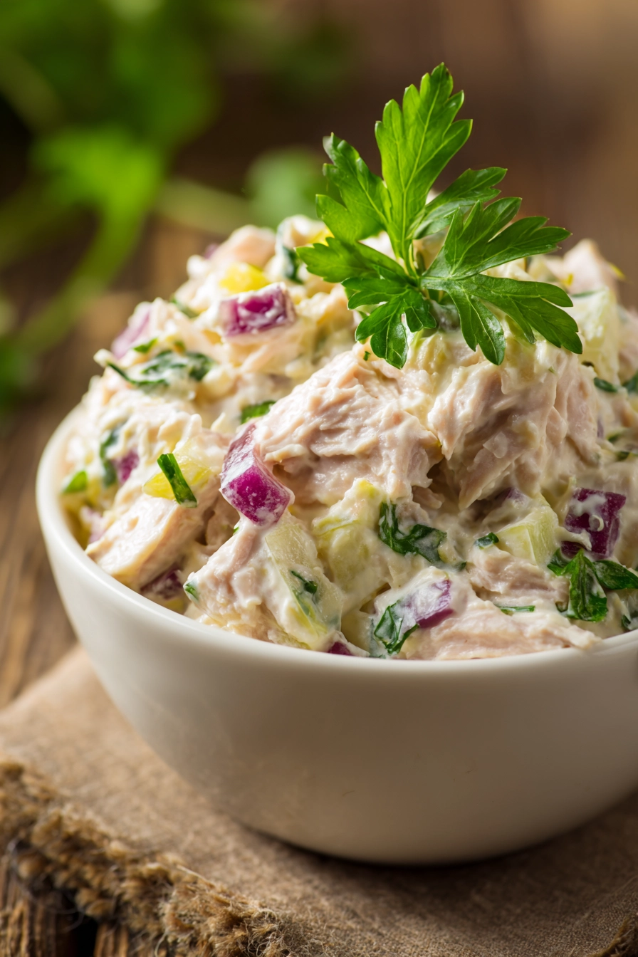 Close-up of classic tuna salad in a shallow white bowl showing creamy texture with chunks of tuna, finely chopped celery and red onion, topped with a sprig of bright green parsley on a rustic wooden surface